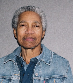 A portrait of Beverly Glenn-Copeland against a gray background, with short gray curly hair, wearing a blue jean collared jacket.