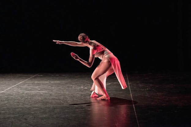 On a dimly-lit stage a performer illuminated by pinkish-red light bends their knees in fourth position and leans forward towards their outstretched arm. 