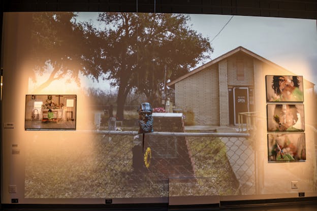 A gallery wall with a picture of Angel Lartigue in front of an altar on the left, a bust of a bearded head in the center, and a vertical row of three pictures of Angel Lartigue holding a medical needle filled with estrogen. Projected onto the wall is an image of a fence surrounding a small cemetery and a building with a sign which reads “Yes, We’re Open.”