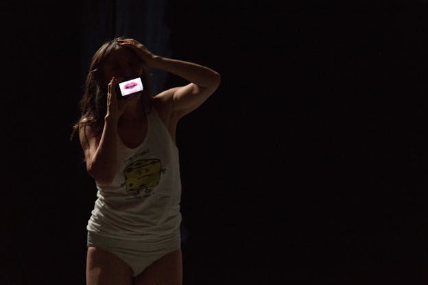 A performer in white underwear and white graphic tank top with a caricature of cheese on it, holds  a small screen showing a mouth in front of their mouth.
