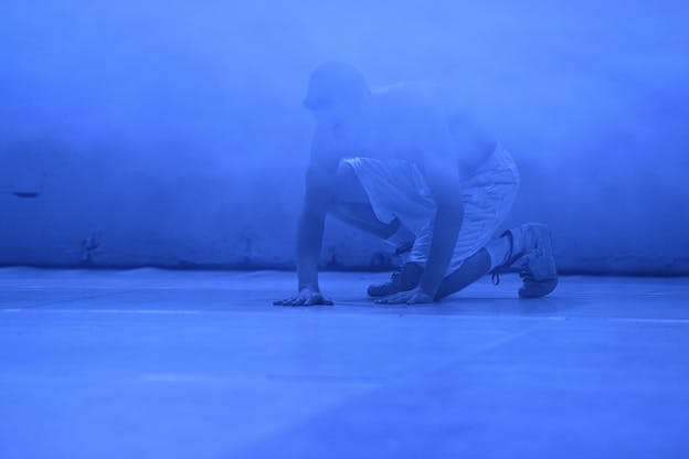 A figure in white shorts and sneakers stand in room lighted in blue with a misty fog surrounding them.