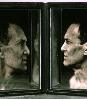 Two framed side angled black and white portraits of William Anastasi are angled towards each other so that they face each other. Each image is partially reflected in the inner edge of the other image. In these portraits, he is depicted with short dark hair and is in front of a dark background. 