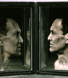Two framed side angled black and white portraits of William Anastasi are angled towards each other so that they face each other. Each image is partially reflected in the inner edge of the other image. In these portraits, he is depicted with short dark hair and is in front of a dark background.