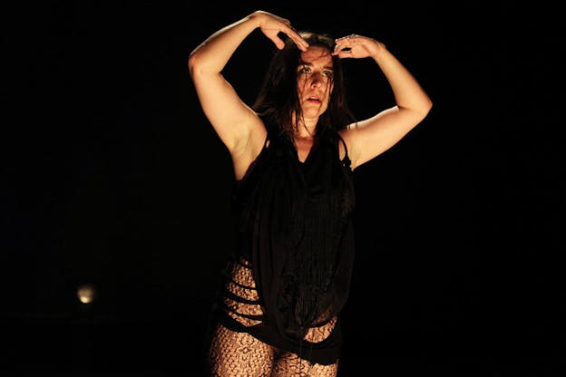 Performer staring offstage, bending their arms over their head, dressed in black clothing and illuminated against a pitch black background. 