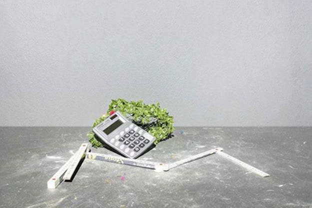 Still-life of a foldable ruler placed on a metal gray table in front of a calculator resting against a clump of green substance. 