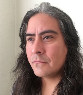 Portrait of Raven Chacon, his head turned at an angle as he stares into the distance. He has long black and grey hair and wears a grey flannel, and stands in front of a cream colored wall.