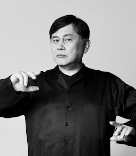 A black and white portrait of Koichi Makigami, who stands in sharp focus before a solid light background. His head is turned slightly to the left while his body faces the camera. His elbows are raised and his hands are posed loosely in front of him with pointer fingers extended. He is wearing a black silk button up top with frog closures. 
