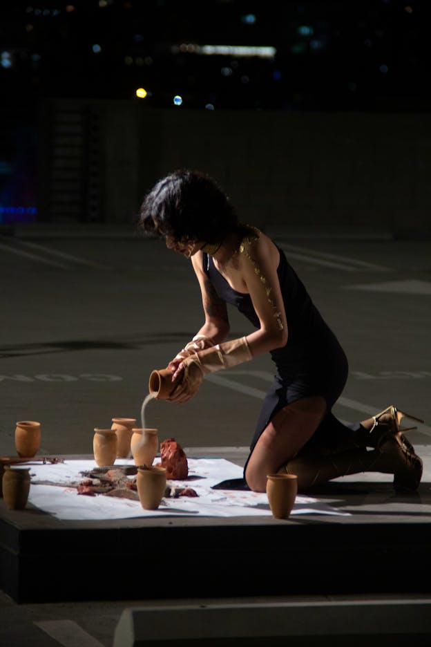 Angel Lartigue kneels in a parking lot, pouring ash or sand from a clay pot onto a large white square covered with several other clay pots and a dissected fish.