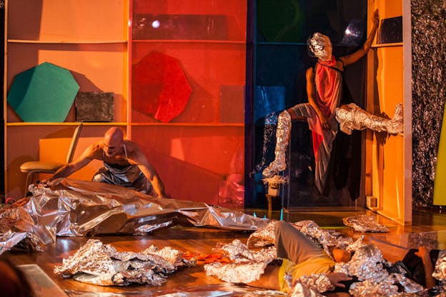 A figure sitting on a high stool is dressed in a black outfit with a red scarf and aluminum foil wrapped around their head and legs. Another figure laying on the floor is covered by foil in a similar way. A third figure is wrapping something in aluminum foil.  