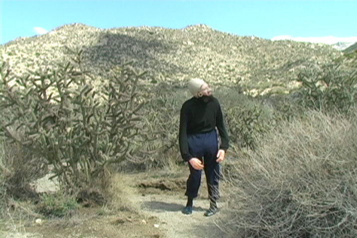 A video still of Ben Tor walking on a path on a mountain, surrounded by shrubbery. She wears a skin colored cap on her head, a beard, and large fake hands and appears mid-stance and peering towards the right.  