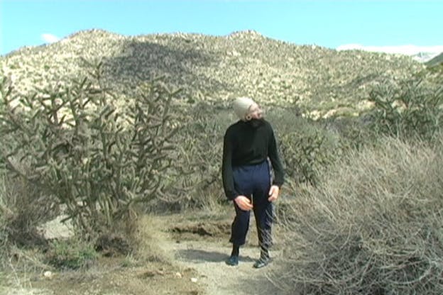 A video still of Ben Tor walking on a path on a mountain, surrounded by shrubbery. She wears a skin colored cap on her head, a beard, and large fake hands and appears mid-stance and peering towards the right.  