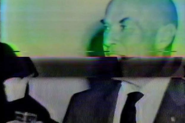 Blurry close-up of a bald person's turned face hued in neon green and blue cut off at the lower lip and divided by a glitchy black strip from their grainy black and white tuxedoed shoulders.