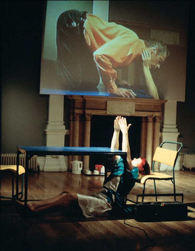 A performance artist lays under a table with their head supported on a chair, their hands lifted upwards. Behind them is a screen projection of a man kneeling down, his had pulled downwards by two extended hands.