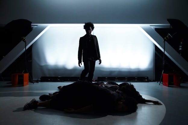 Performer, shadowed and dressed all in black, stands in a dimly lit room in front of an LED projector screen looking down at a row of shadowed bodies lying face-down on the floor.
