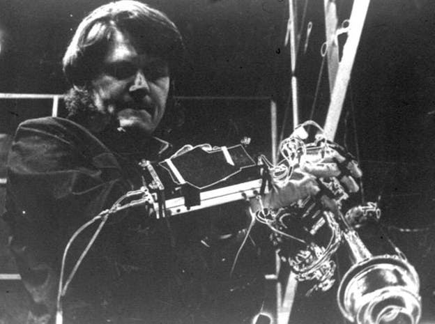 A close up black and white image of Gordon Mumma in front of sound ewquipment in a dark space. He wears a dark shirt and has a concentrated expression on his face. 