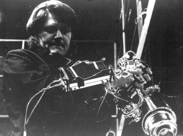 A close up black and white image of Gordon Mumma in front of sound ewquipment in a dark space. He wears a dark shirt and has a concentrated expression on his face. 
