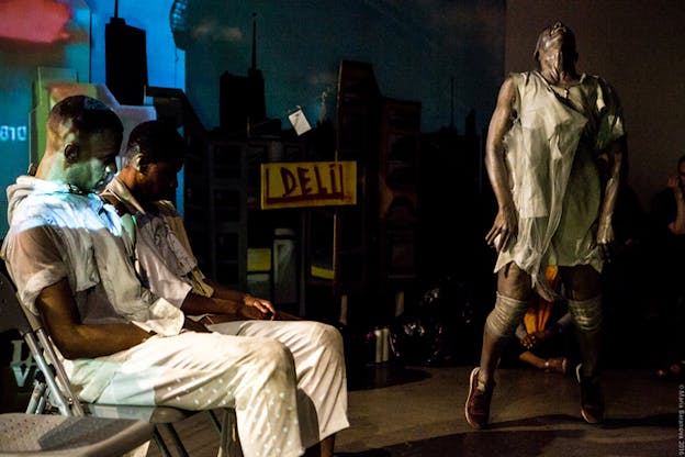 Two perfromers sit on chairs next to each other while a third in disheveled white clothing stands on his toes with his head angled back screaming. Between them a yellow sign with the words 