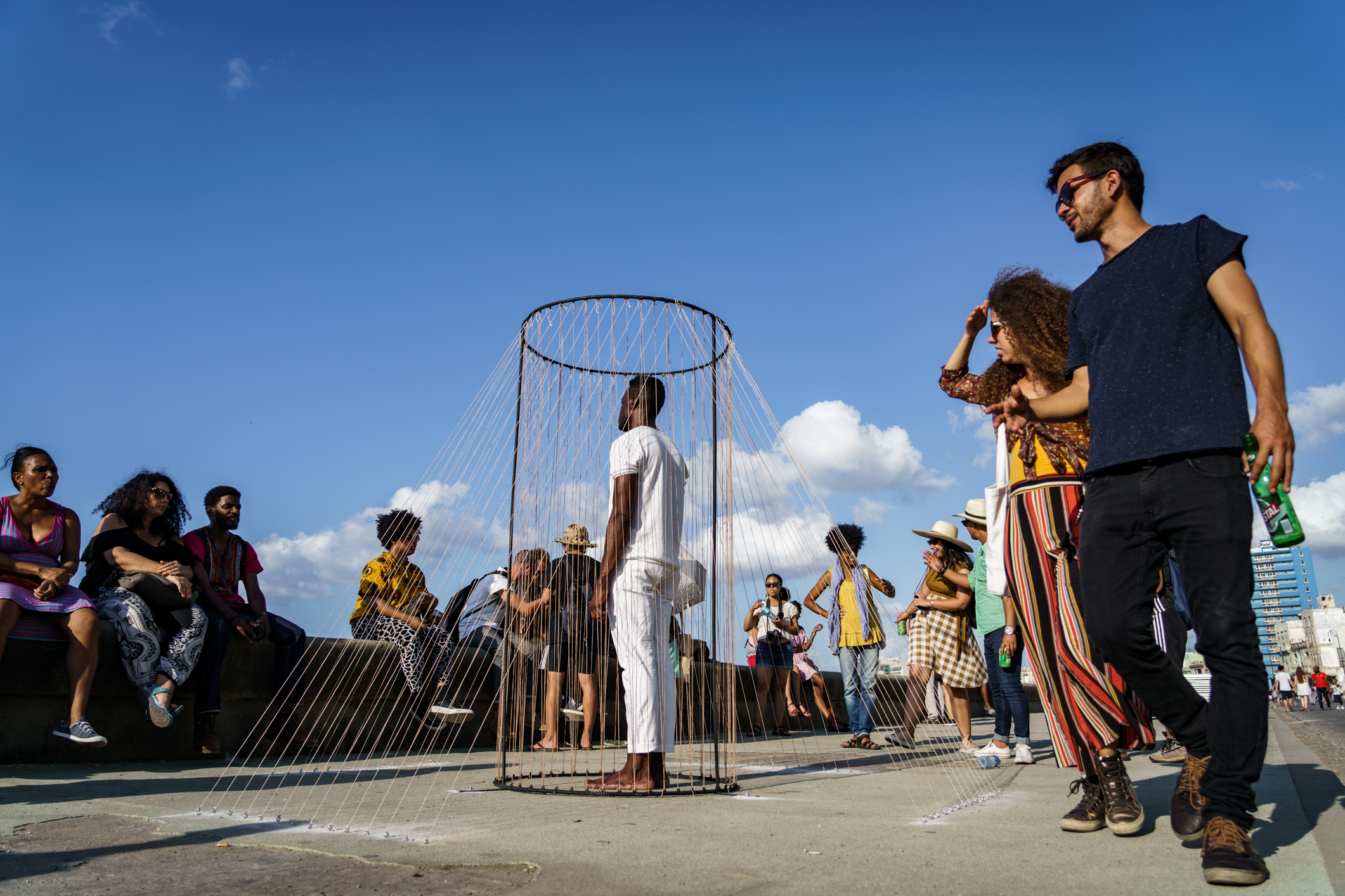 A performance still of Carlos Martiel wearing all white standing in a metal cylindrical frame with numerous metal wires eminating from it, connecting from the top of the frame to the ground around it. This is set outside, a top pavement. Audience members sit on a bench directly facing Martiel and some passberys glance one. The sky above them all is bright blue and slightly cloudy. 