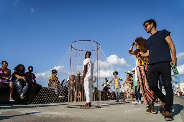 A performance still of Carlos Martiel wearing all white standing in a metal cylindrical frame with numerous metal wires eminating from it, connecting from the top of the frame to the ground around it. This is set outside, a top pavement. Audience members sit on a bench directly facing Martiel and some passberys glance one. The sky above them all is bright blue and slightly cloudy. 