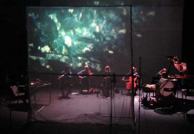 A small orchestra performs on a dimly lit stage in between two screens projecting shards of turquoise resembling light filtering through water. 