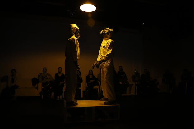 Two performers stand in a dark room under a hanging light. Only their silhouettes are illuminated. They wear gas masks and full hazmat suits.
