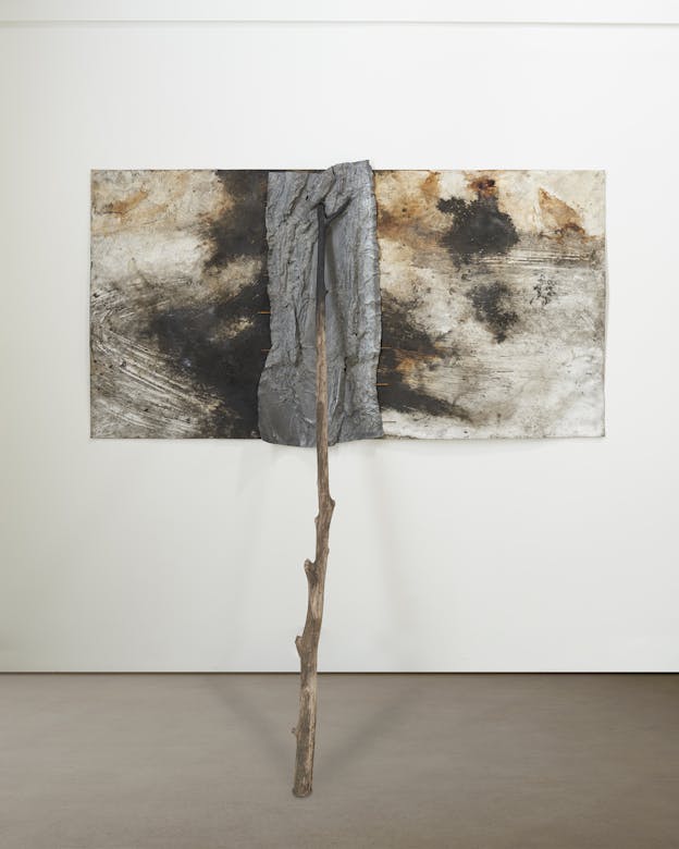 A long rectangular multimedia painting with black ink and soil on a white sheet of paper. A thick, crinkled grey strip vertically bisects the painting in imperfect halves, fastened to the paper using copper-colored wire. A long stick with an ink covered end rests against the painting in the middle of the grey strip, extending to the floor.