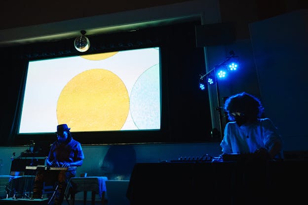 Newman Taylor Baker sits masked, playing the washboard in a dark space in front of a large screen lit up with yellow and light blue circles. To his left is Lori Scacco wearing reflective sunglasses and a mask in front of a mixing board.