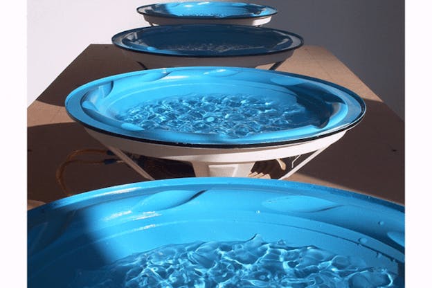 An installation image of four blue plates on white stands held above speakers. A shallow layer of water ripples within the plates. 