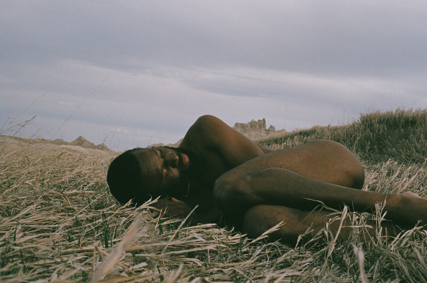 A video still of JJJJJerome Ellis laying in a field of wheat, naked and curled in a fetal position. His eyes are closed. Behind him, some tan colored stone structures are visible. 
