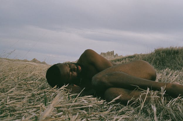 A video still of JJJJJerome Ellis laying in a field of wheat, naked and curled in a fetal position. His eyes are closed. Behind him, some tan colored stone structures are visible. 