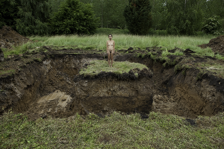 A naked figure facing the viewer stands in the center of a plain, its surrounding dug out leaving them on a island patch of land.