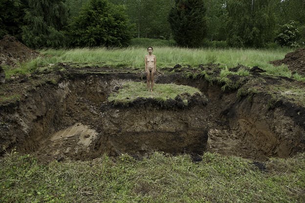 A naked figure facing the viewer stands in the center of a plain, its surrounding dug out leaving them on a island patch of land.