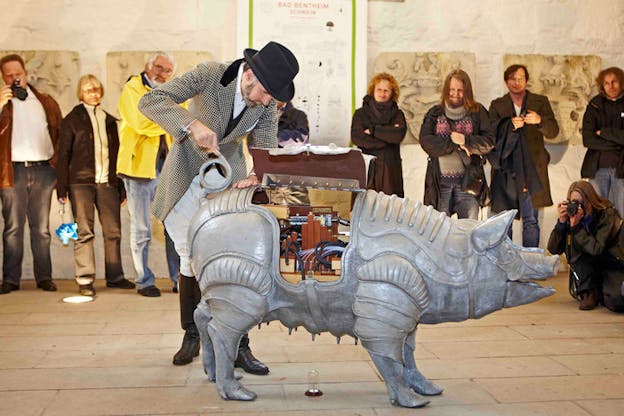 An image of Lincoln performing with the mechanical pig, cranking the pigs tail. Lincoln wears thigh high black boots, white pants, a white and black checkered coat, and a black fedora. Behind him, an audience watches from the walls of the chapel.