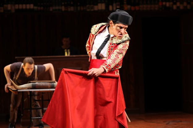 A performance still of a performer dressed in a red and gold bullfighter's outfit with a bruise on their cheek. They hold a piece of bright red cloth and stare intently towards the distance. Behind them, another performer holds themselves over a table with a bull's horns on it. This performer in the back and the shelf of wine bottles behind them are all blurred.