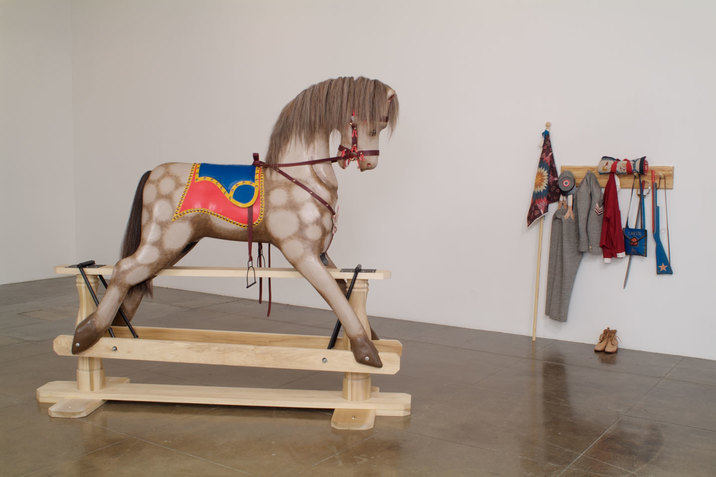An installation image of a grey spotted horse wearing a bridle and red and blue saddle, elevated on wooden planks. On the white wall behind this, there is a shelf with military clothing, shoes, a plastic blue gun, and a decorative quilted flag. 