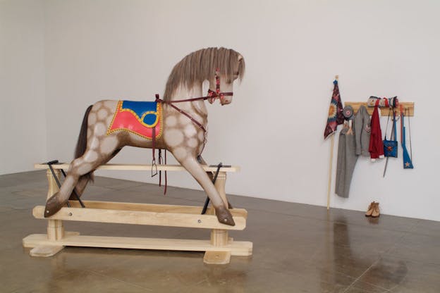 An installation image of a grey spotted horse wearing a bridle and red and blue saddle, elevated on wooden planks. On the white wall behind this, there is a shelf with military clothing, shoes, a plastic blue gun, and a decorative quilted flag. 