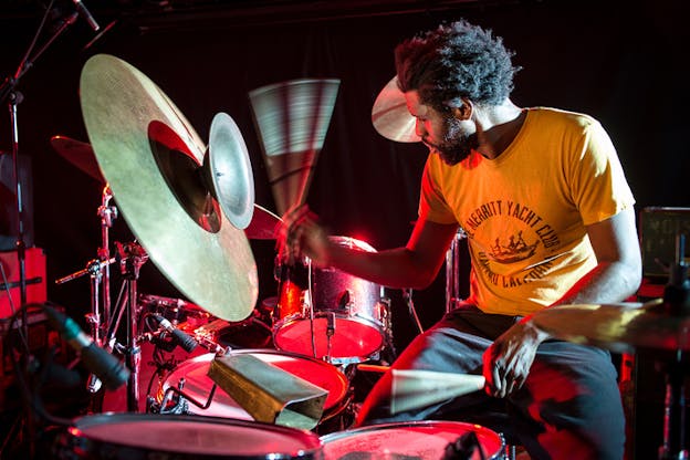 Close up shot of a seated figure in black sweatpants and orange t-shirt behind drums and other percussion instruments playing them while illuminated by a red light.