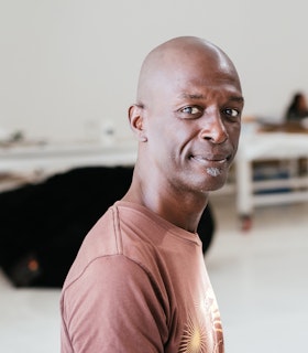 A portrait of David Thomson wearing a light red t-shirt with a yellow sunburst design. He is bald and has a small grey gotee. The background of the image depicts blurred white shelves. 