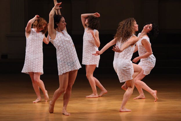 Performers in white short dressed with flower cut details move on a wooden floor.