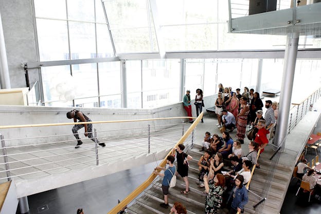 People down on a stairwell watch as a performer climbs stairs with their body facing downwards and balloons flowing upwards connected to them with a string surrounding the neck.