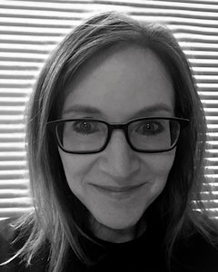 Black and white portrait of Kathleen Flynn smiling towards the camera, dressed in a dark shirt and thick framed glasses, with shoulder-length straight hair parted to the side.