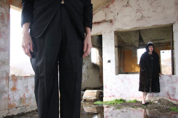 Two performers stand in a room covered in peeling white paint. On the left, only the black pant legs and arms of one person are seen as they stand close to the camera. On the right, there is a person wearing a thick fur coat and black stilettos and leaning against an opening in the wall behind them. The floor appears slightly wet and some weeds and dirt cover it. 