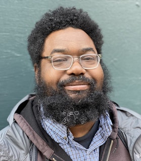 Mike Cloud smiles softly into the camera. He wears glasses and a collared button down shirt under a windbreaker.