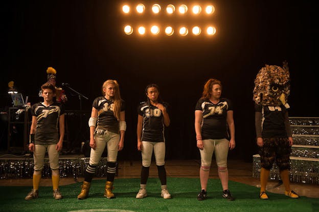 Four performers dressed in football players clothing, with a fifth person wearing an owl mascot mask. 