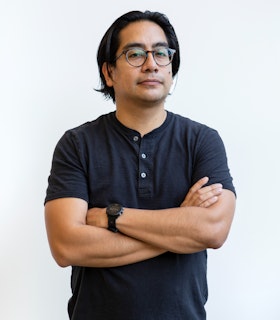 Ronny Quevedo stands with his arms crossed before a white background, facing the camera. He wears round, tortoiseshell glasses, a short sleeve, asphalt-colored henley, black jeans, and a black, round-faced digital watch on his left wrist.