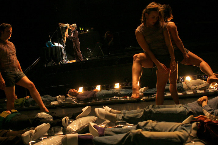 Several people lay on the floor, wearing blue jeans and white slippers. Three people wearing short tunics step over the laying people. In the far background, a person is seen wearing all black and playing an instrument.
