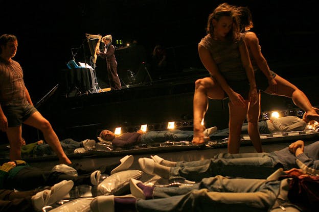 Several people lay on the floor, wearing blue jeans and white slippers. Three people wearing short tunics step over the laying people. In the far background, a person is seen wearing all black and playing an instrument.