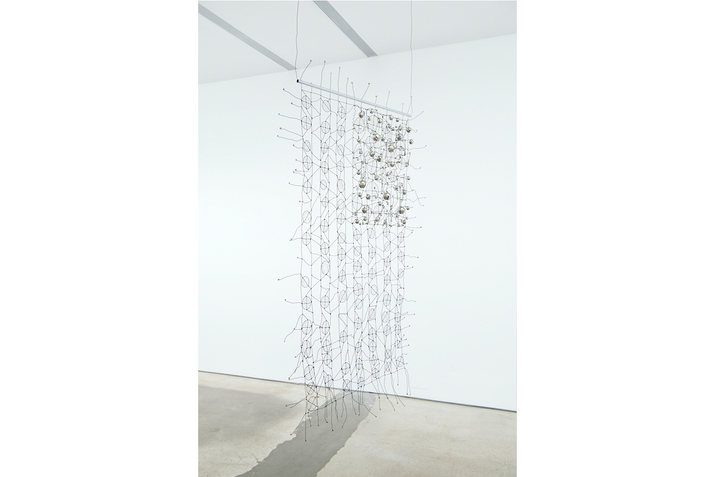 A rectangle shaped wire weaved structure hangs from a ceiling. Divisions of stripes exist in it, with seven of them weaved in circles and six in waves. On the top left corner another rectangle is densely weaved, forming the U.S flag.