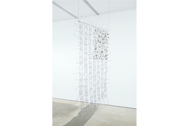 A rectangle shaped wire weaved structure hangs from a ceiling. Divisions of stripes exist in it, with seven of them weaved in circles and six in waves. On the top left corner another rectangle is densely weaved, forming the U.S flag.