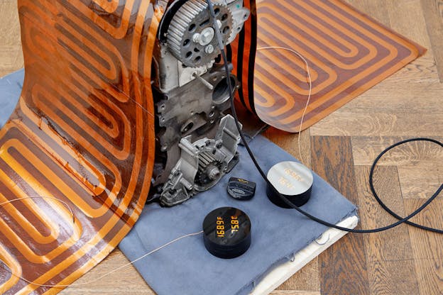 A metallic mechanisms underneath a orange and maroon glass carpet. In its front two black circular devices with numbers of Fahrenheit and Celsius.